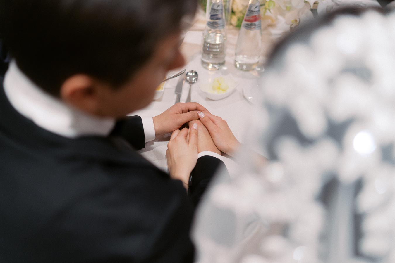 Wedding couple holding hands at a table, showcasing intimate connection and love. Table setting includes cutlery and bottled water, suggesting a fine dining experience.