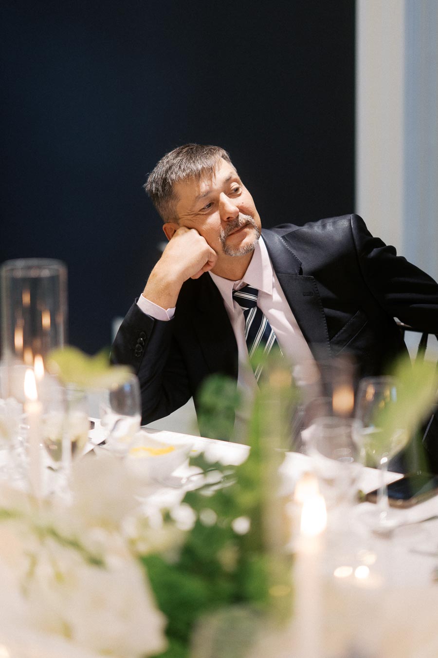 A man in a suit sits at an elegantly set dinner table with candles and floral arrangements, appearing thoughtful and relaxed.