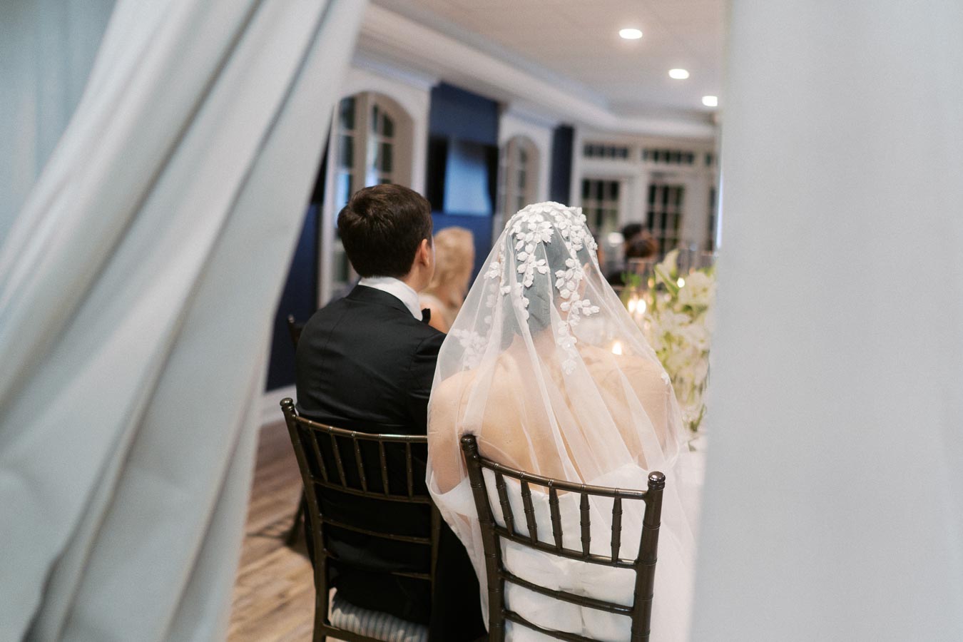 Rear view of a bride and groom sitting at a wedding reception, with the bride wearing a floral veil and the groom in a black suit, captured in an elegantly decorated venue.