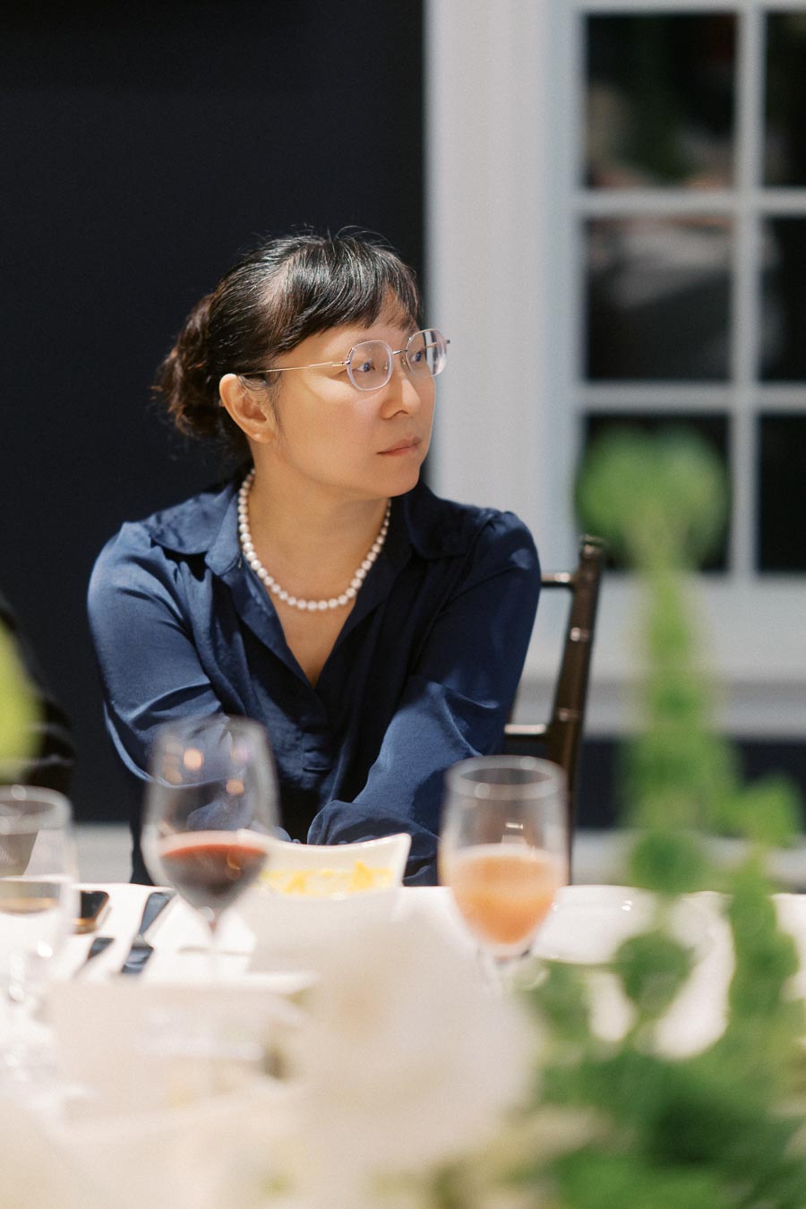 A woman in a blue blouse and glasses, wearing a pearl necklace, seated at a dinner table with glasses of wine and blurred greenery in the foreground.