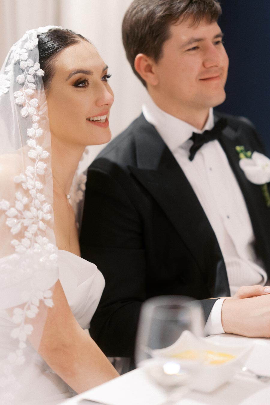 Elegant bride and groom seated together at a wedding reception, bride wearing a floral lace veil and groom in a classic black tuxedo, both smiling.