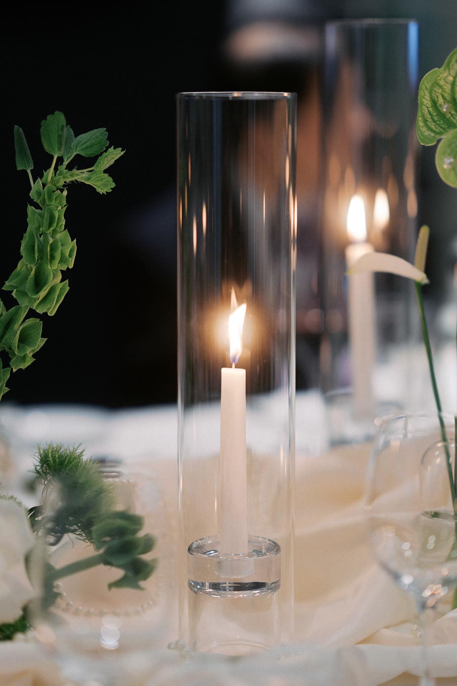 Elegant dinner table setting with lit candles in tall glass holders, surrounded by green foliage and delicate flowers, creating a romantic ambiance.
