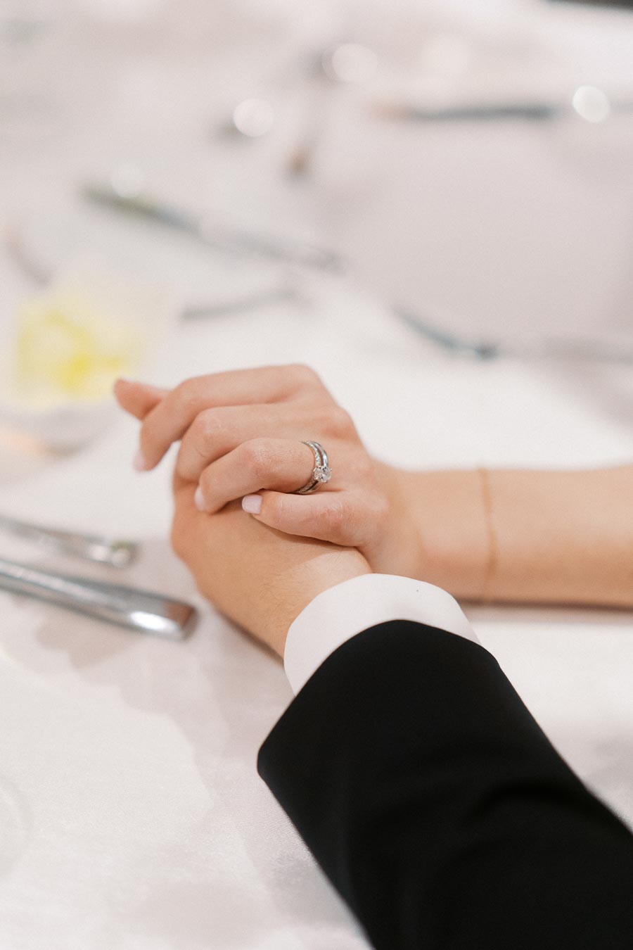 Close-up of a couple holding hands at a wedding, focusing on an elegant engagement ring on the woman's finger, with a blurred background of a formal table setting.
