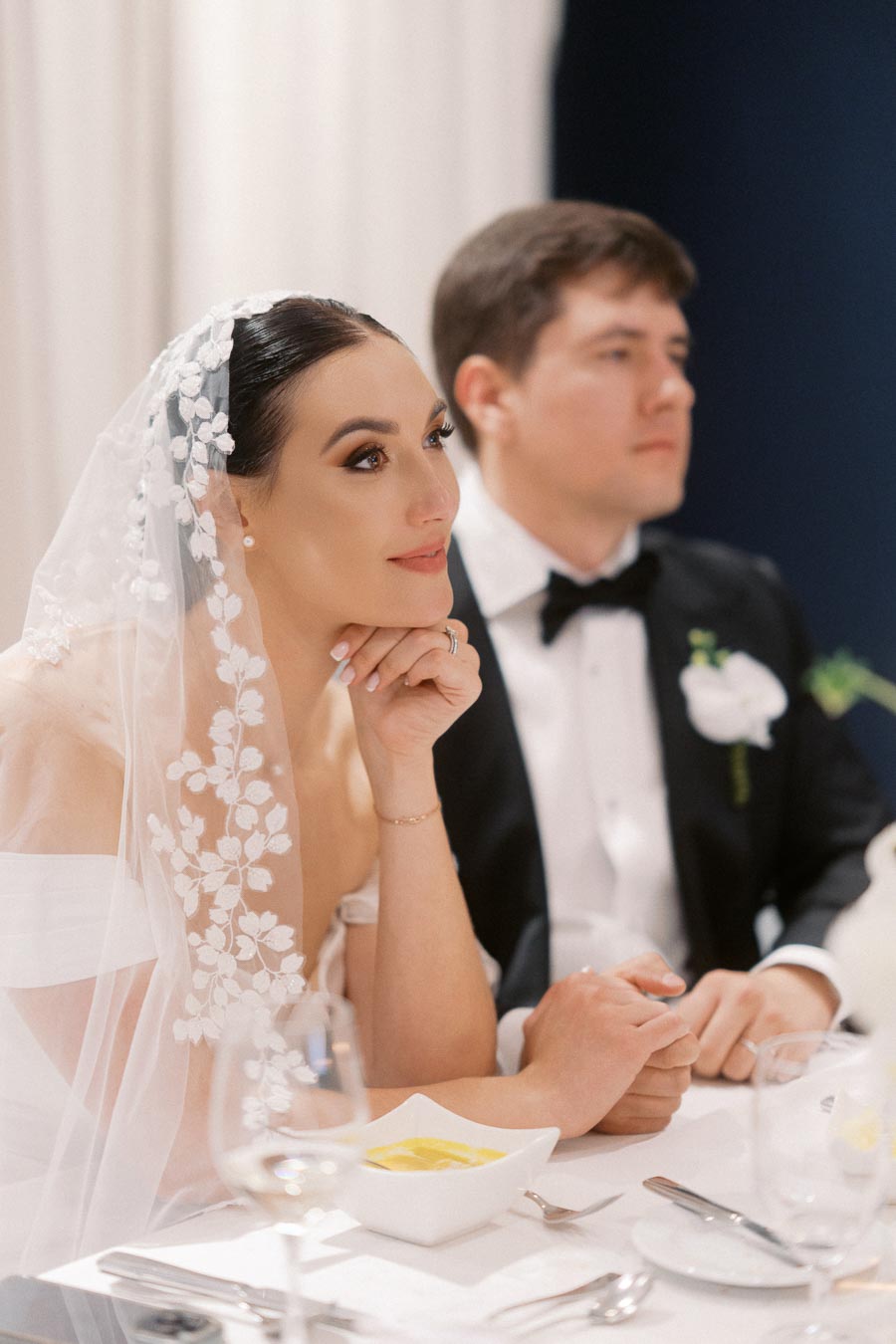 A bride in an elegant lace veil and off-the-shoulder gown sits thoughtfully at a wedding reception table next to a groom in a black tuxedo with a boutonniere, highlighting an intimate wedding moment.