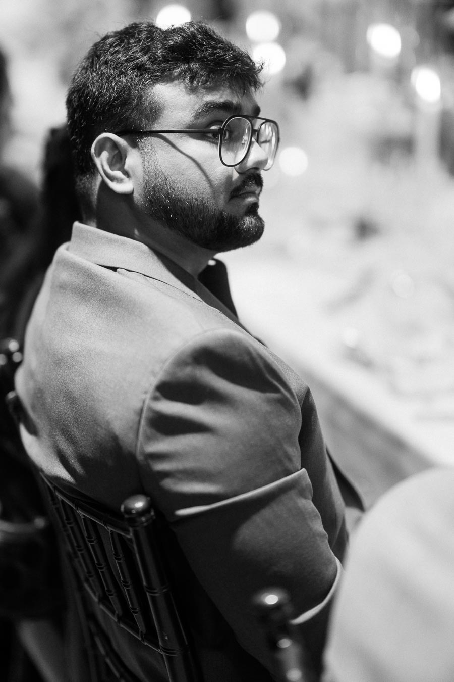 Black and white portrait of a man with glasses, sitting at an event, wearing a suit jacket.