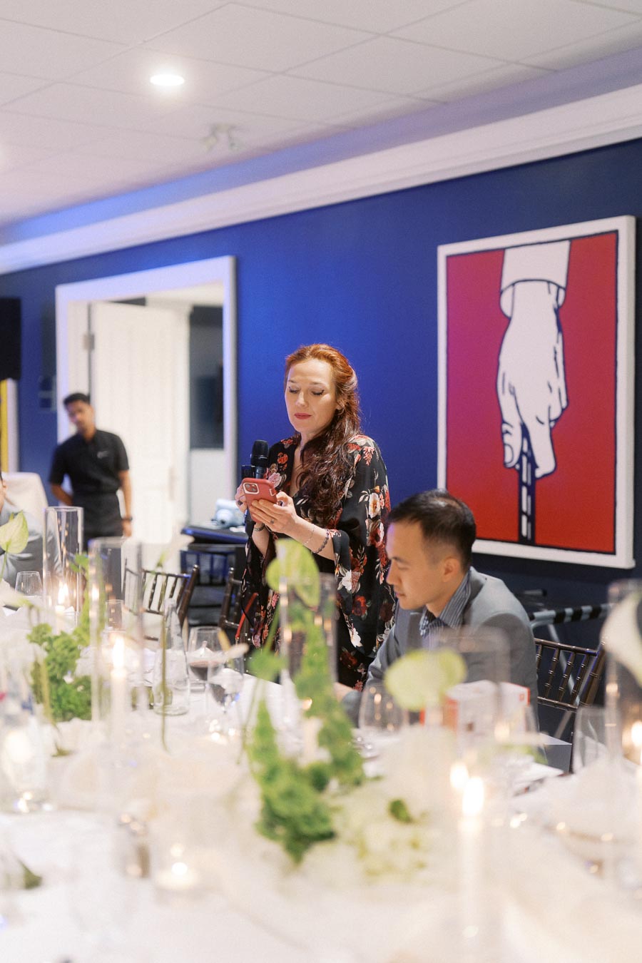 Woman giving a speech at a dinner event, holding a microphone and phone, with elegantly set table and decorative centerpiece in the foreground, and modern artwork on a blue wall in the background.