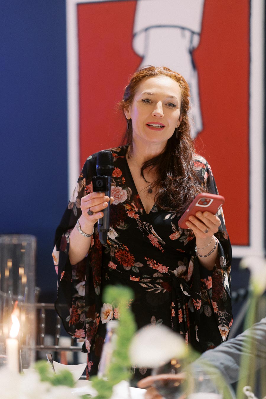 A woman in a floral dress holding a microphone and smartphone, speaking at an indoor event with a modern art backdrop.