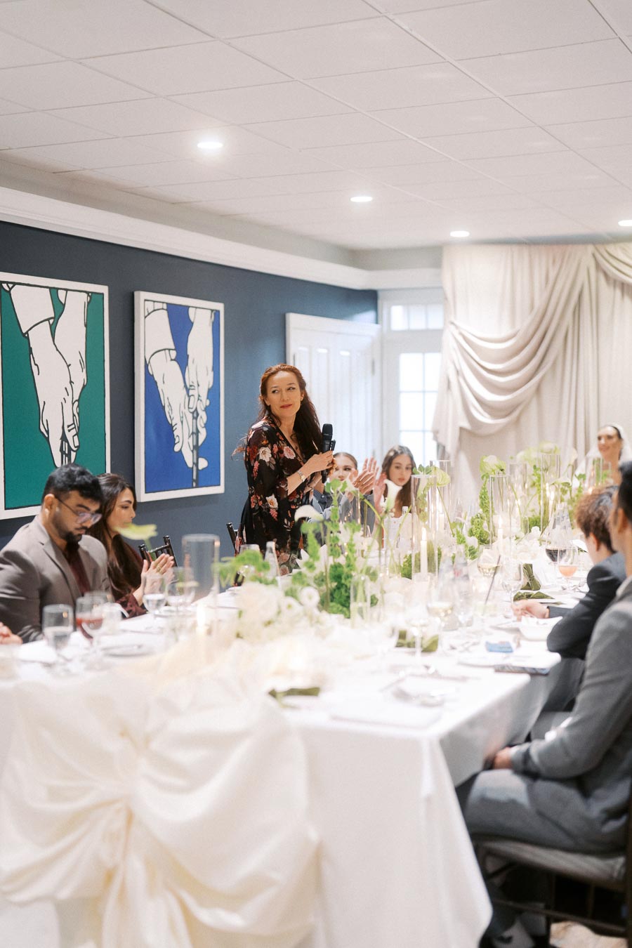 Elegant indoor dinner event with a woman giving a speech at a beautifully decorated table, featuring green floral arrangements and white drapery, attended by seated guests in formal attire.