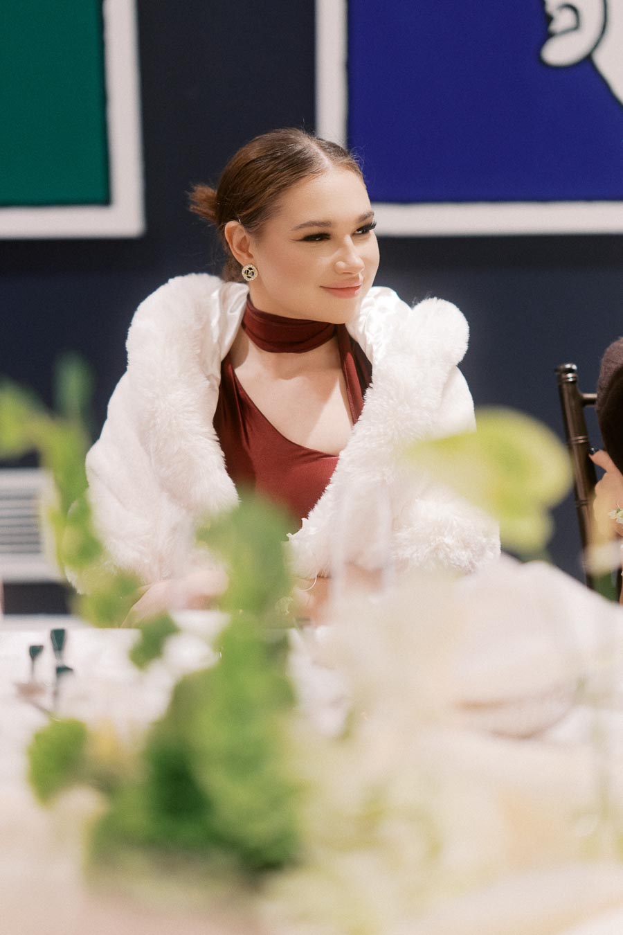 Young woman in elegant attire and white fur shawl sitting at an elegantly set table, with blurred greenery in the foreground and abstract art in the background.