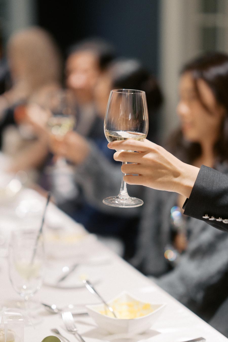 A person holding a wine glass in a celebratory toast at a formal dining table, surrounded by blurred figures in elegant attire.
