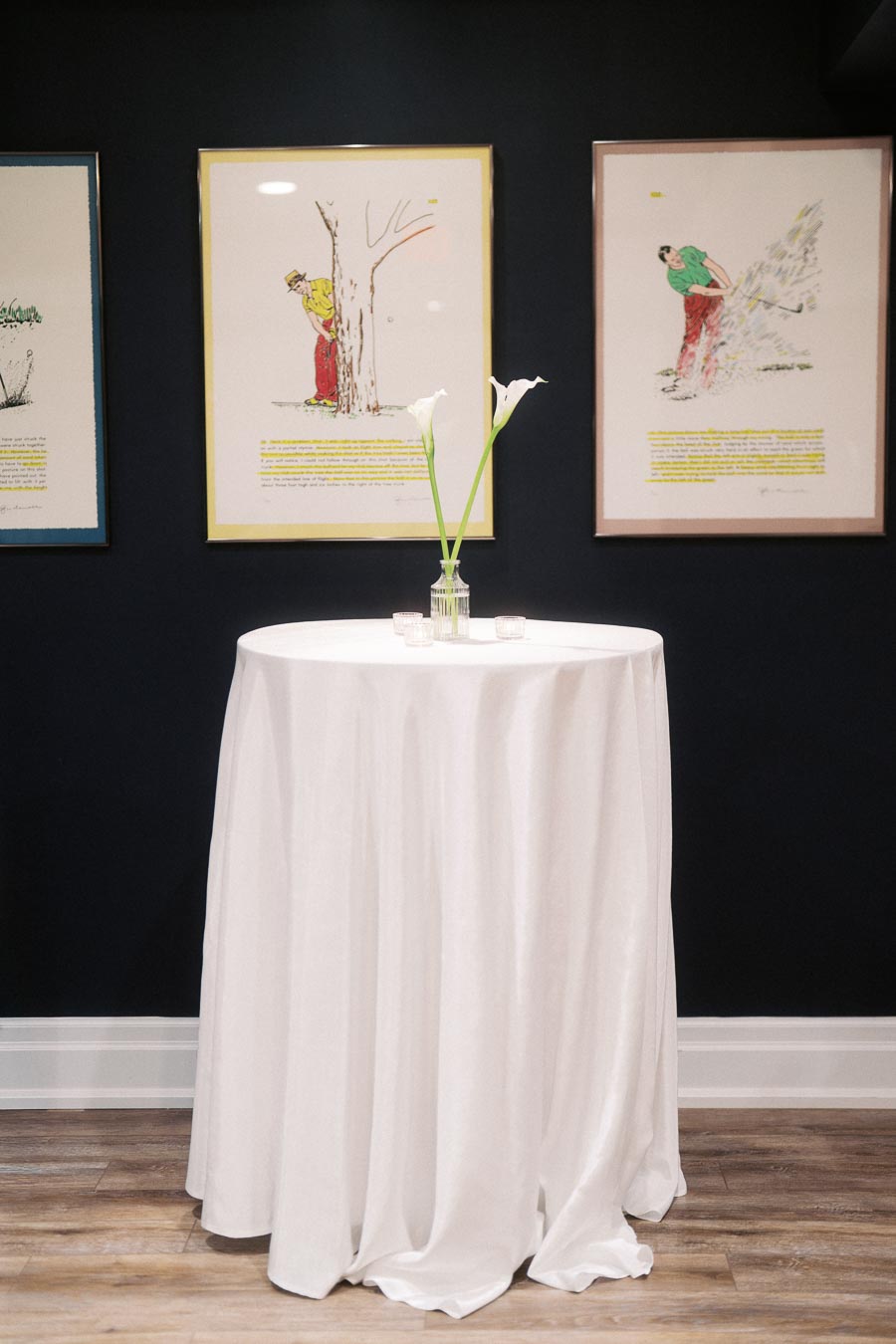 Elegant white-draped cocktail table with two calla lilies in a glass vase, set against a dark wall with framed art prints in a modern gallery space.