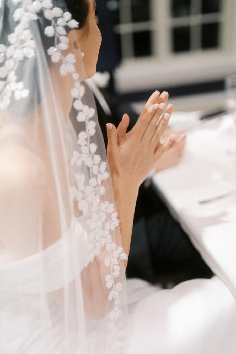 Elegant bride in lace veil clapping hands at wedding ceremony, showcasing intricate floral embroidery and serene atmosphere.