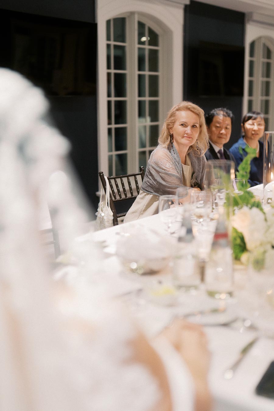 Elegant dinner setting with guests in formal attire, featuring a woman wrapped in a shawl at a beautifully set table with floral centerpieces and soft candlelight.