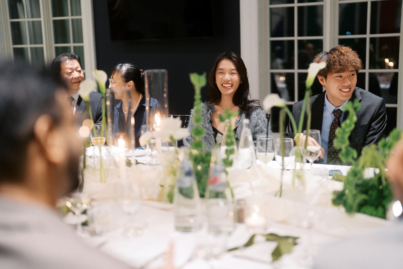 A group of people smiling and enjoying a formal dinner party, seated around a beautifully decorated table with white flowers and candles, creating an elegant and joyful atmosphere.