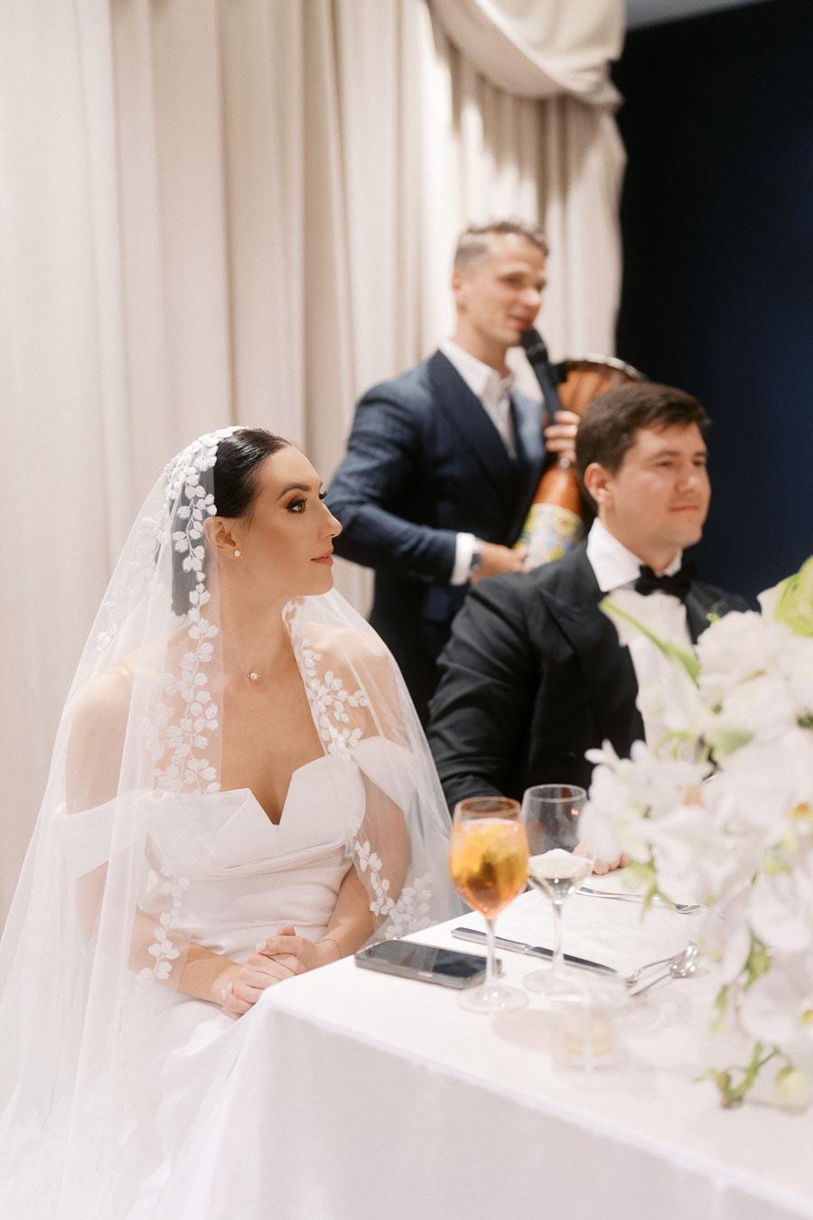 Bride and groom seated at a wedding reception table adorned with white flowers, as a musician plays nearby, creating an elegant and joyful ambiance.