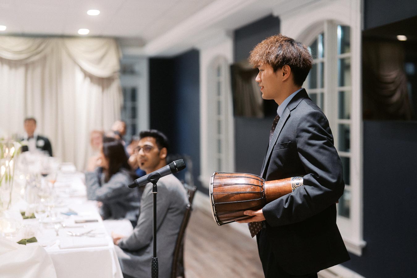 A person in a suit holds a drum while standing near a microphone at an elegant indoor event with seated guests.