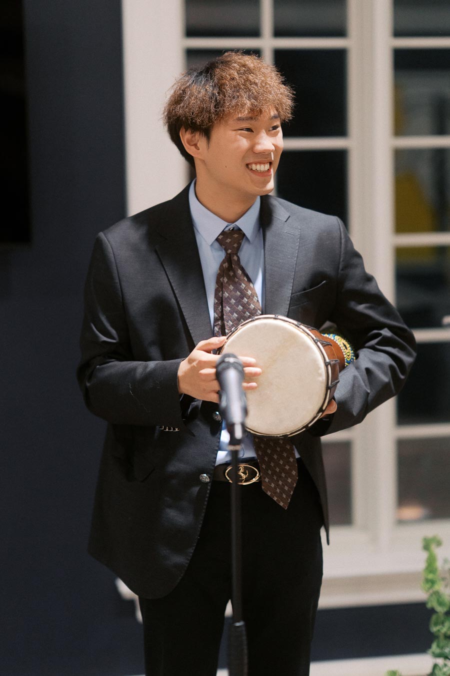 A person in a suit smiling and holding a drum, standing near a microphone in a well-lit room.