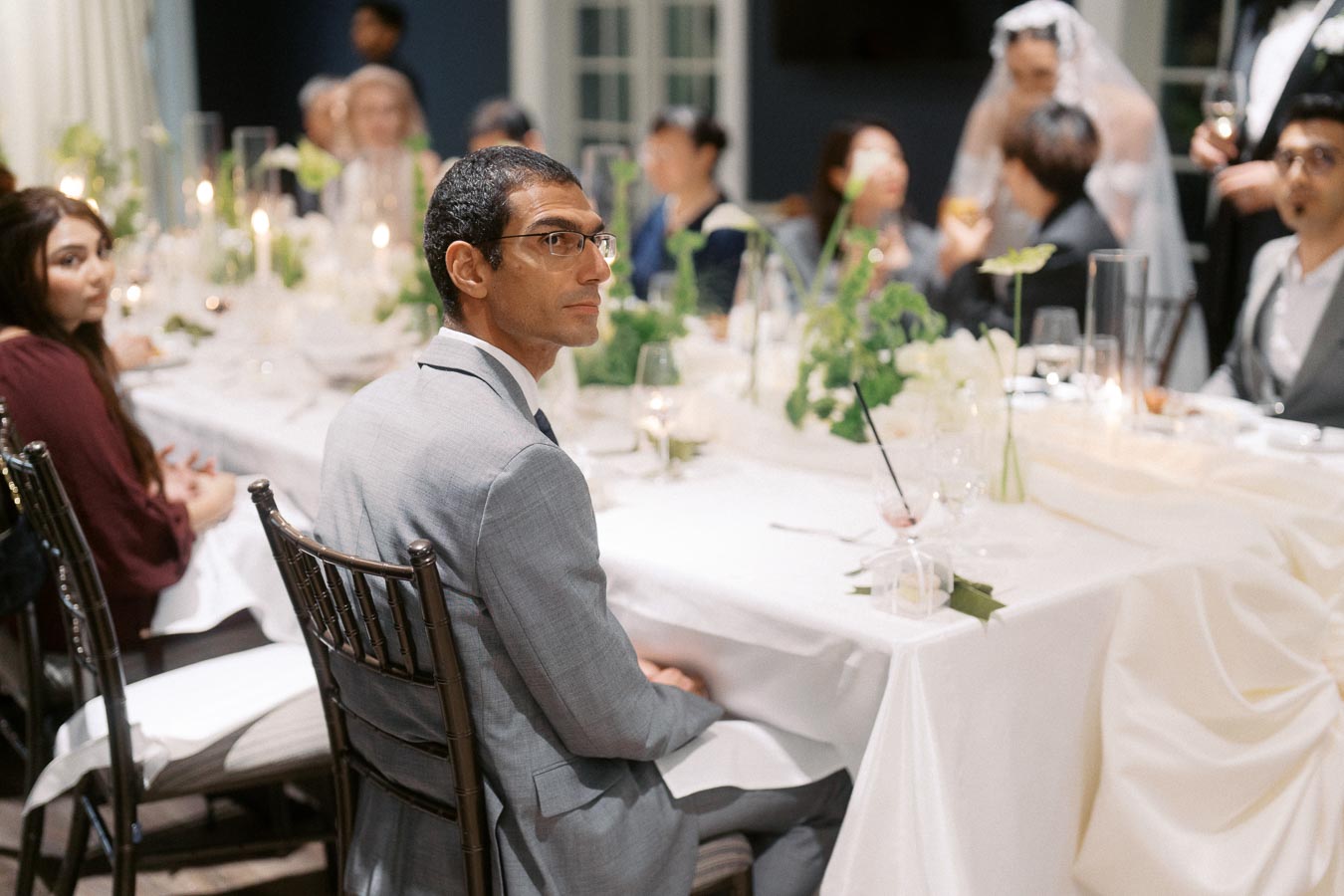 A man in a gray suit sits at an elegantly decorated dining table during a wedding reception. The scene features white tablecloths, green floral arrangements, and guests engaged in conversation, with a bride in the background. The atmosphere is formal and celebratory.