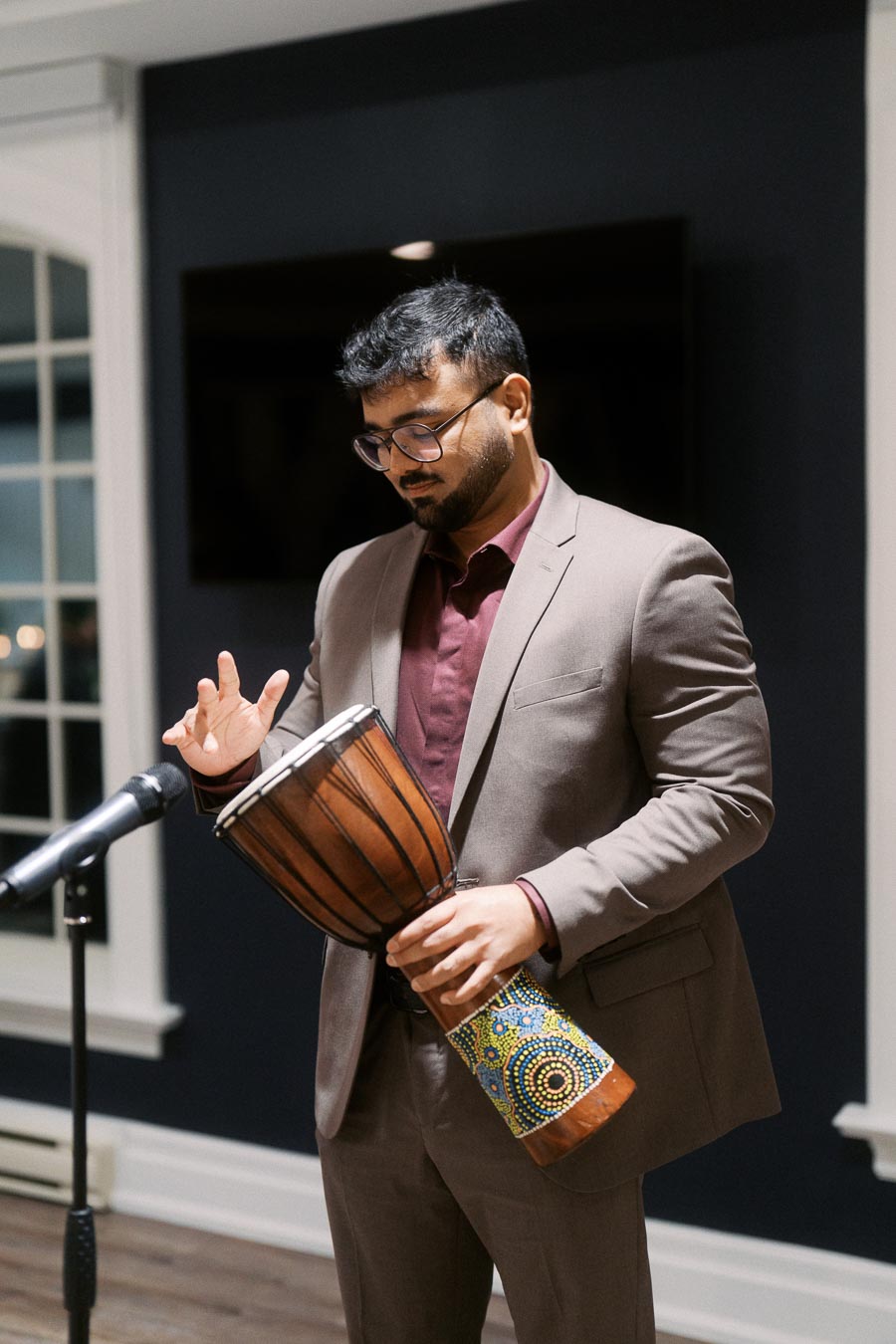 Man in a suit playing a djembe drum by a microphone in a dimly lit room.
