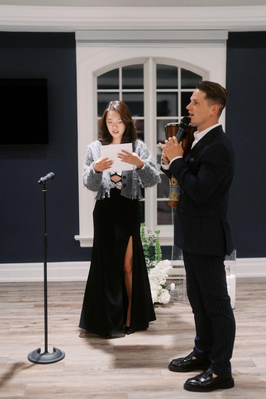 A woman in a formal black dress reads from a sheet of paper while a man in a suit holds a microphone and a percussion instrument indoors. The setting is elegant, featuring a large window and wooden flooring.