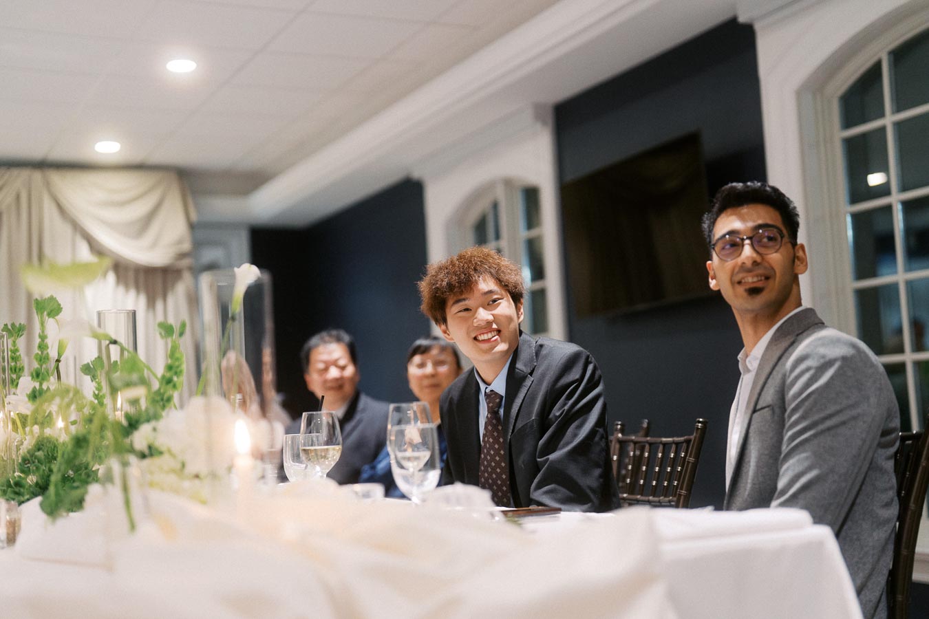 A group of people in formal attire smiling at a table set with glassware and floral arrangements during an elegant indoor event.