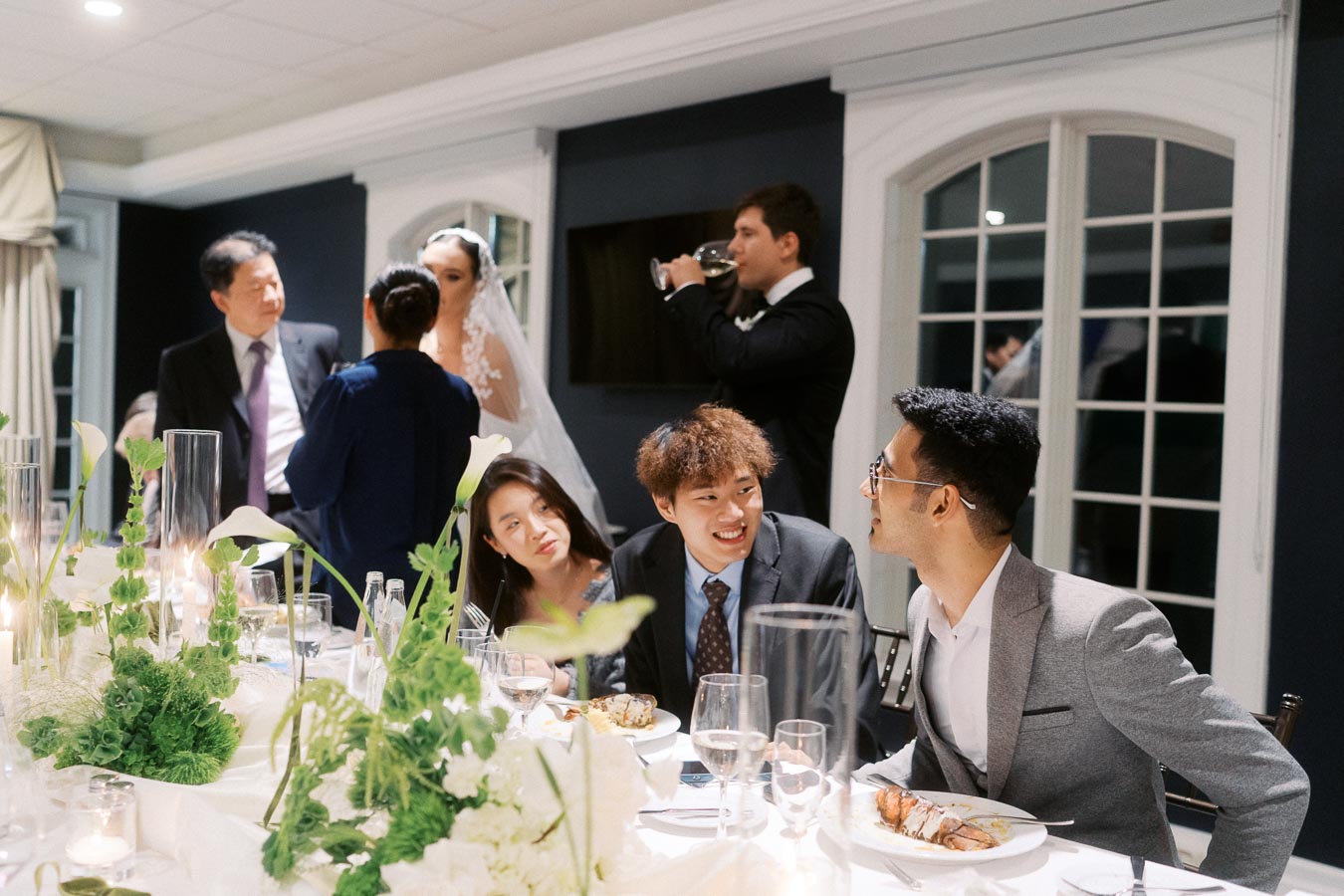 A group of well-dressed wedding guests enjoying a festive dinner at a beautifully decorated reception table, featuring elegant green and white floral centerpieces and soft candlelight.