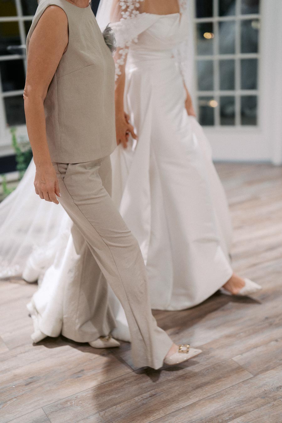 Elegant bridal and formal attire captured during a wedding event, featuring a flowing white wedding dress with intricate lace details and a stylish beige pantsuit, set against a background of large windows and wooden flooring.