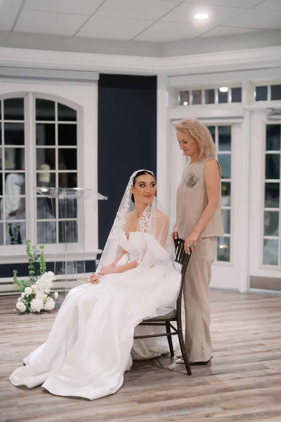 Bride in elegant white gown and veil sits on a chair, accompanied by a woman in beige attire in a bright room with large windows, floral decorations, and wooden flooring.