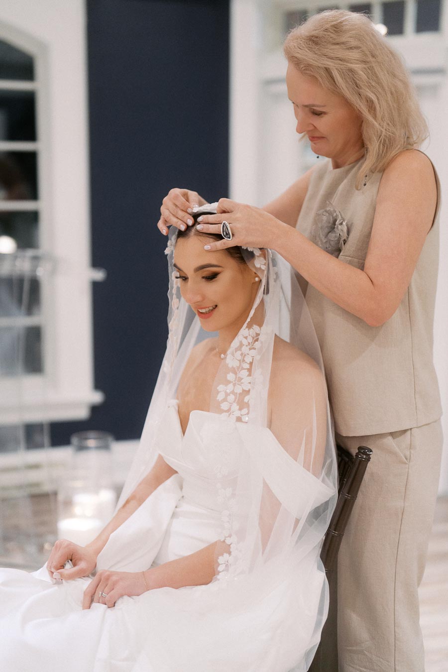 Bride getting ready with help from a woman adjusting her floral lace veil, smiling and seated indoors, wearing an elegant white wedding dress.