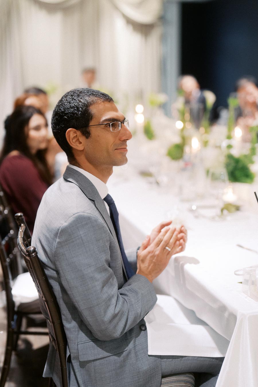 A man in a gray suit and glasses sits at an elegantly set dining table, appearing engaged and thoughtful, with blurred guests and soft lighting in the background.