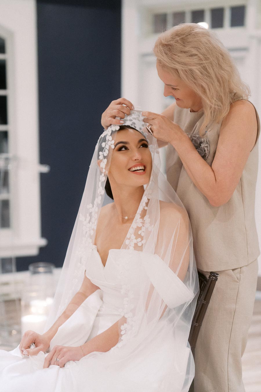 Bride smiling as her veil is adjusted by a woman in a soft, elegant setting.