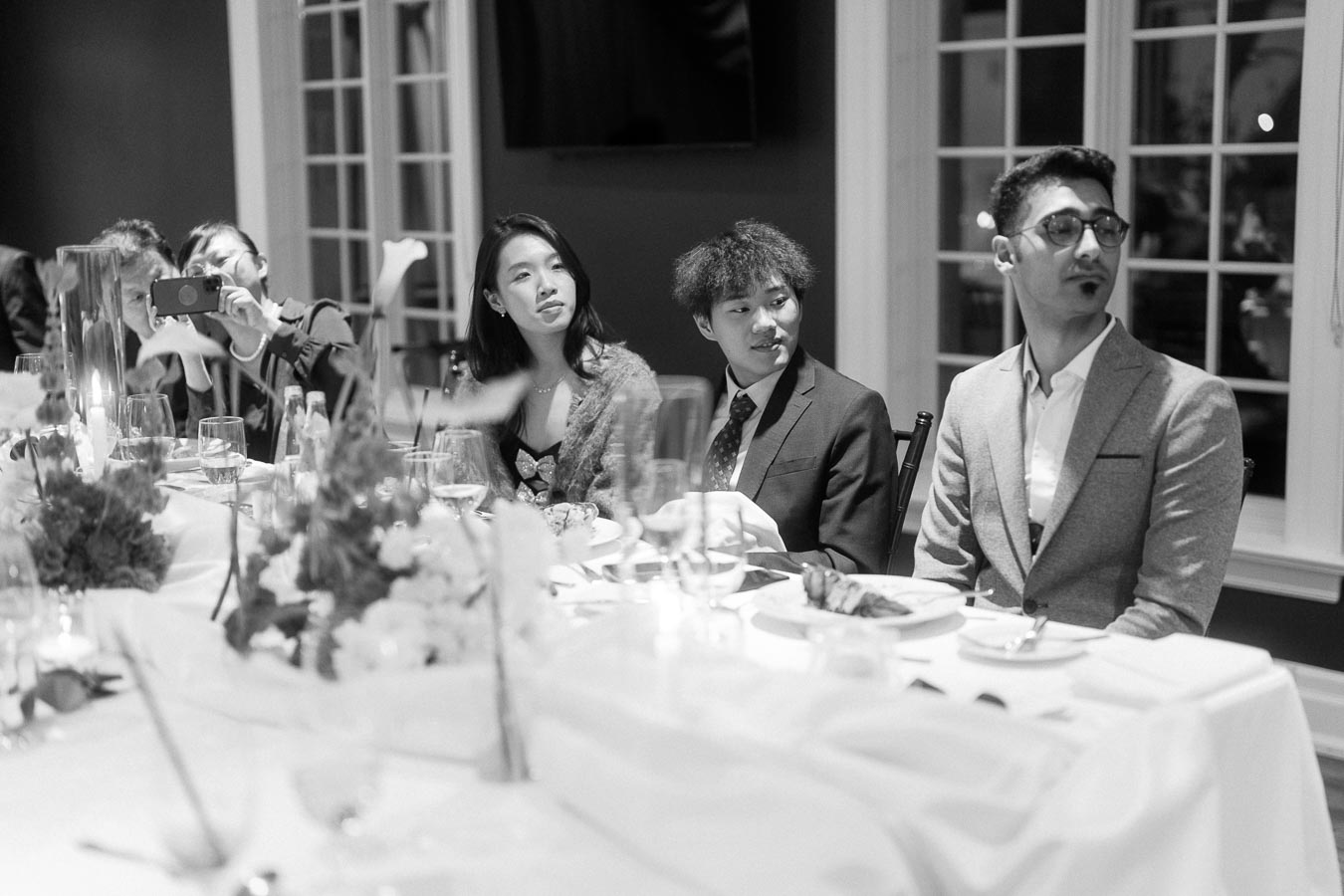 A black-and-white image of a formal dining event with elegantly dressed individuals seated at a table. The setting includes decorative floral arrangements and glasses on a white tablecloth. Attendees appear engaged and attentive, creating a sophisticated and refined atmosphere.