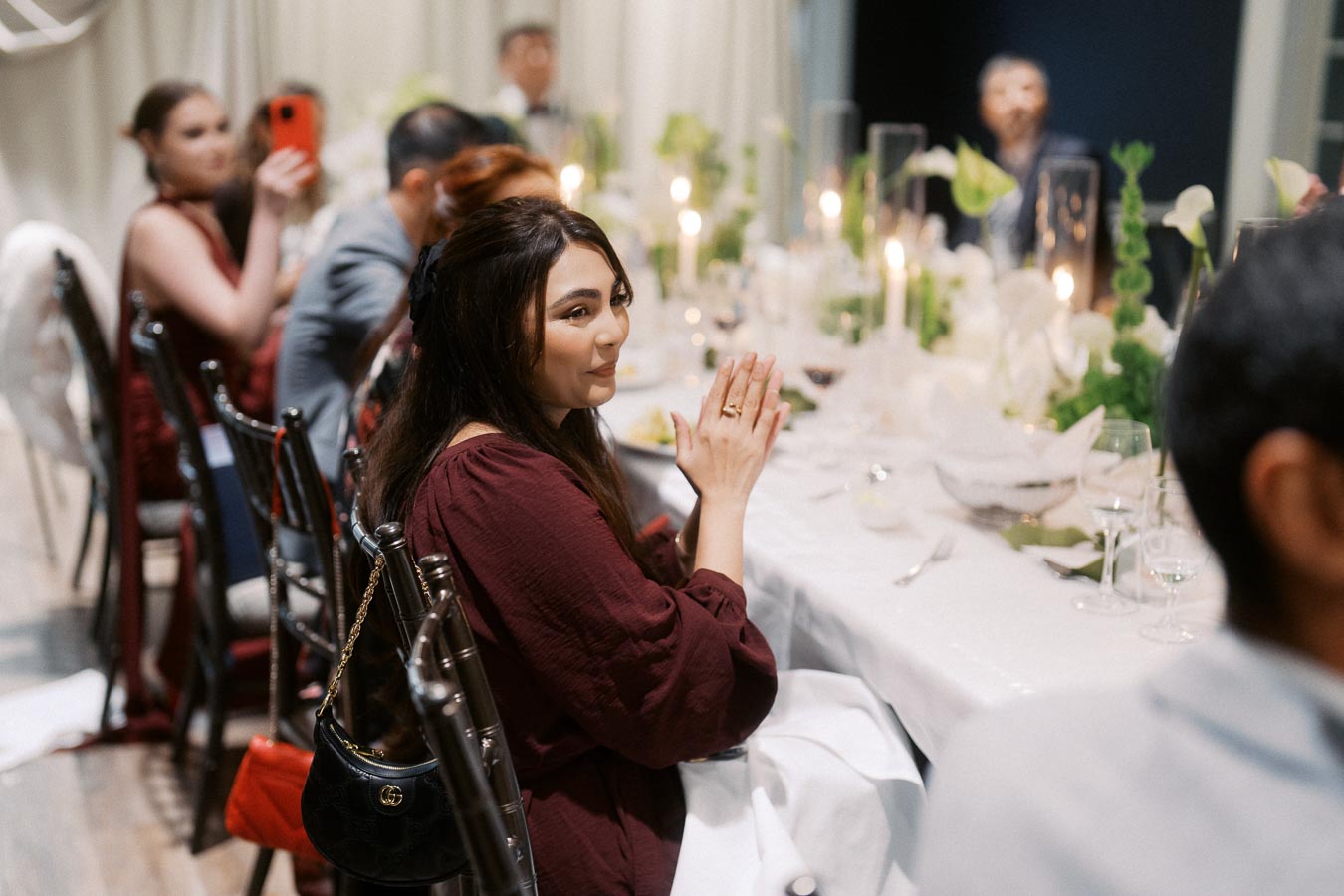 A woman in a maroon dress clapping at a formal event, seated at a beautifully set dinner table with candles and greenery, with other guests in the background.