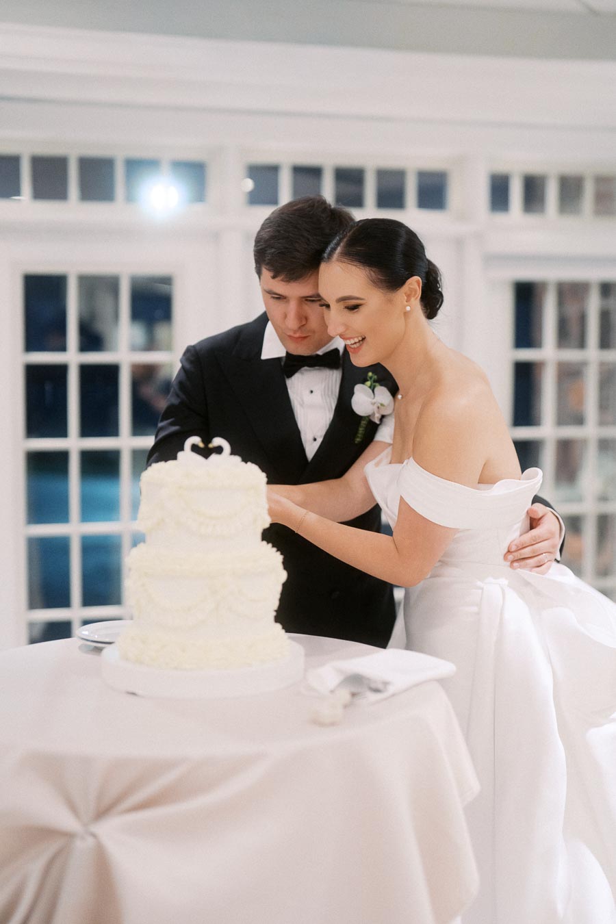 Bride and groom cutting a white tiered wedding cake together in an elegant setting, with the groom wearing a tuxedo and the bride in a strapless white gown.