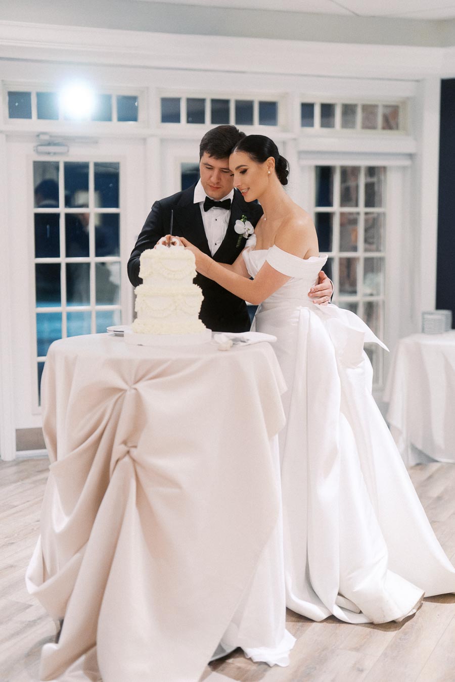 A bride and groom cutting a tiered white wedding cake together in an elegant setting with large windows in the background, both dressed in formal attire.