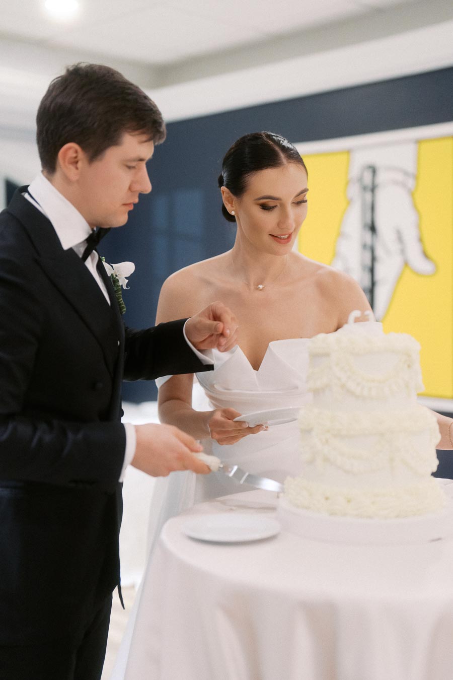 Bride and groom cutting an elegant white wedding cake at their reception, both smiling and dressed in formal attire, showcasing a joyous wedding moment.