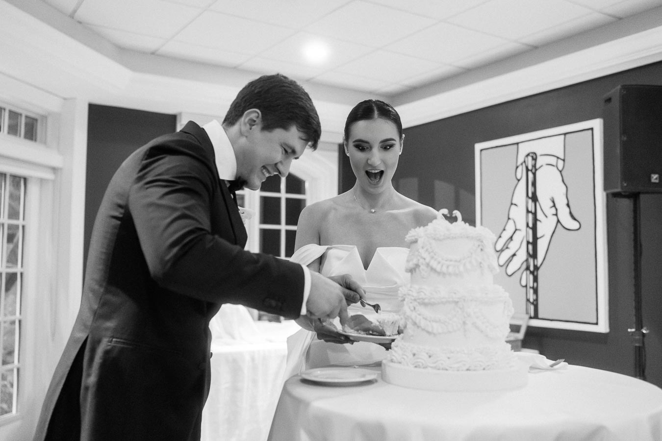 Bride and groom cutting a tiered wedding cake during their reception, with the bride looking surprised and the groom focused on serving a slice. Black and white photograph capturing a joyful wedding moment.