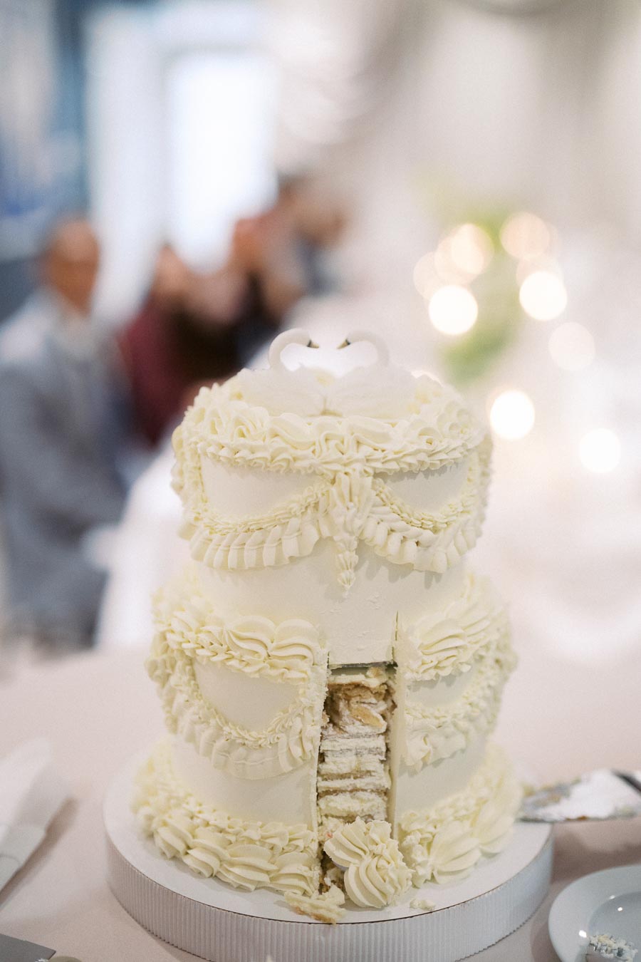 Two-tiered white wedding cake with intricate icing details, partially sliced to reveal a creamy interior, set against a blurred background of elegantly decorated event setting.