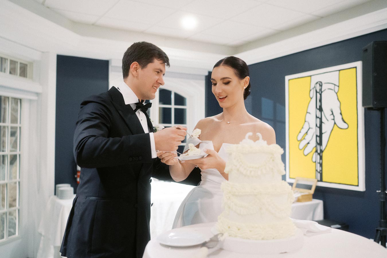A bride and groom joyfully sharing cake at their wedding reception, with a beautifully decorated white cake in the foreground and a contemporary painting on the wall behind them.