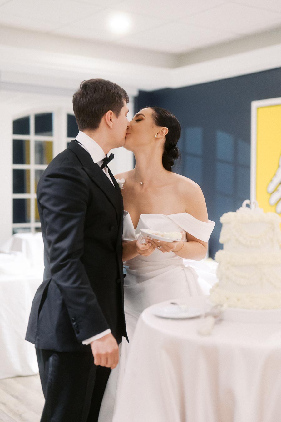 Bride and groom sharing a kiss at their wedding reception, dressed in formal attire, next to a decorative white wedding cake on a table.