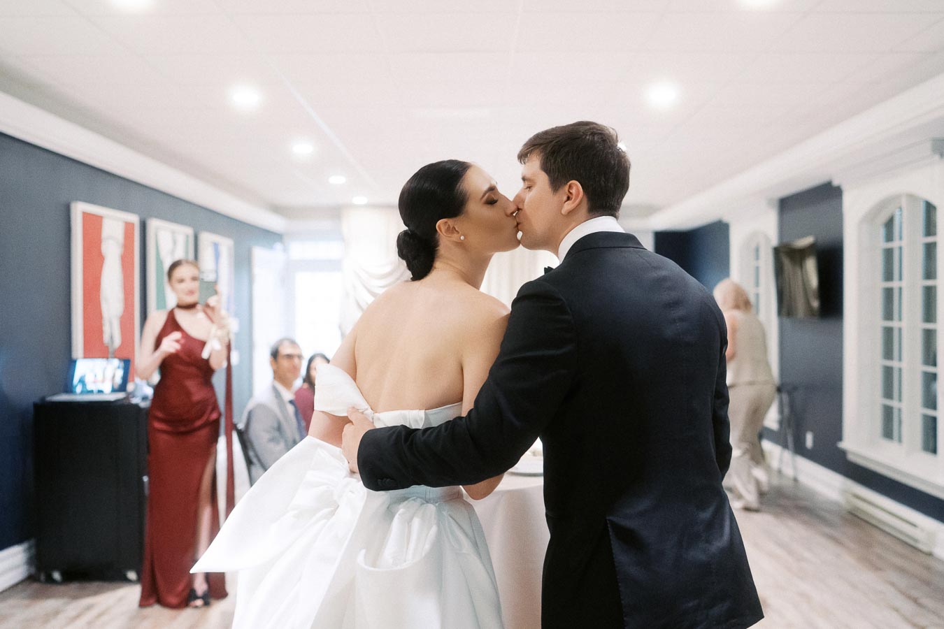 Bride and groom sharing a kiss at their wedding reception, with elegantly dressed guests celebrating in a beautifully decorated room.