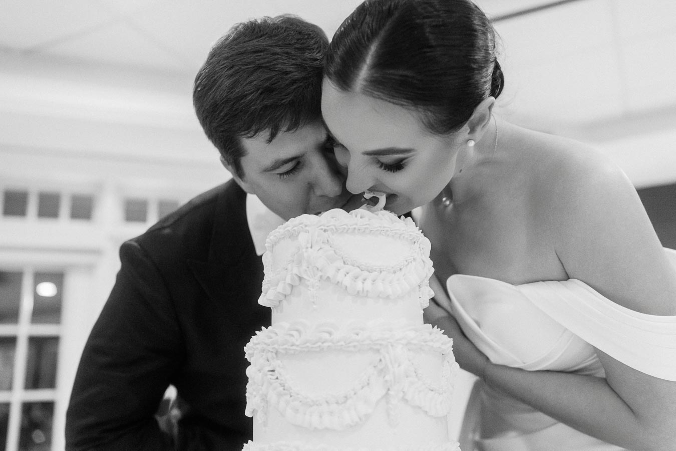 Black and white photo of a bride and groom playfully biting into a multi-tiered wedding cake, capturing a joyful moment during their celebration.