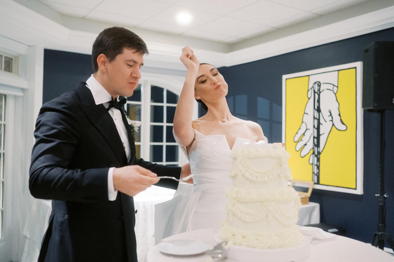 A bride playfully gestures while slicing a tiered wedding cake with the groom beside her, both in formal attire, in a decorated indoor venue with artwork in the background.