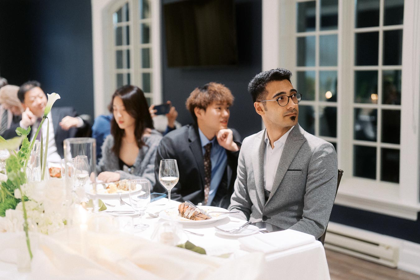A group of elegantly dressed individuals seated at a banquet table during a formal dinner event, featuring decorative plants and glassware, with large windows in the background.