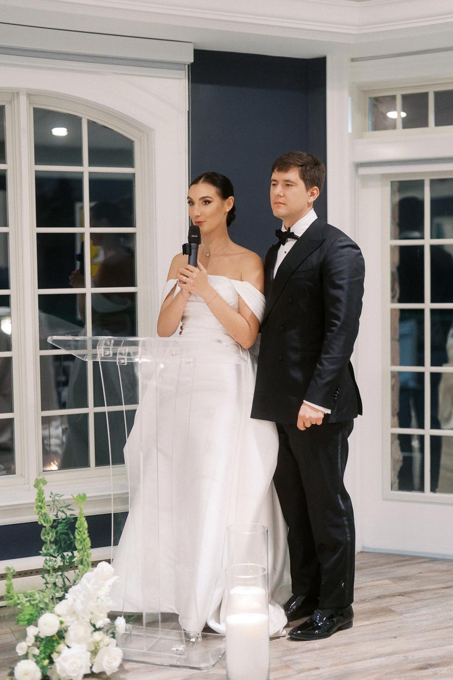 Elegant couple in formal attire standing together at a wedding reception, with the woman holding a microphone. The bride is wearing a sophisticated white gown and the groom is in a classic black tuxedo. They're positioned near a transparent lectern with floral decorations and candles.