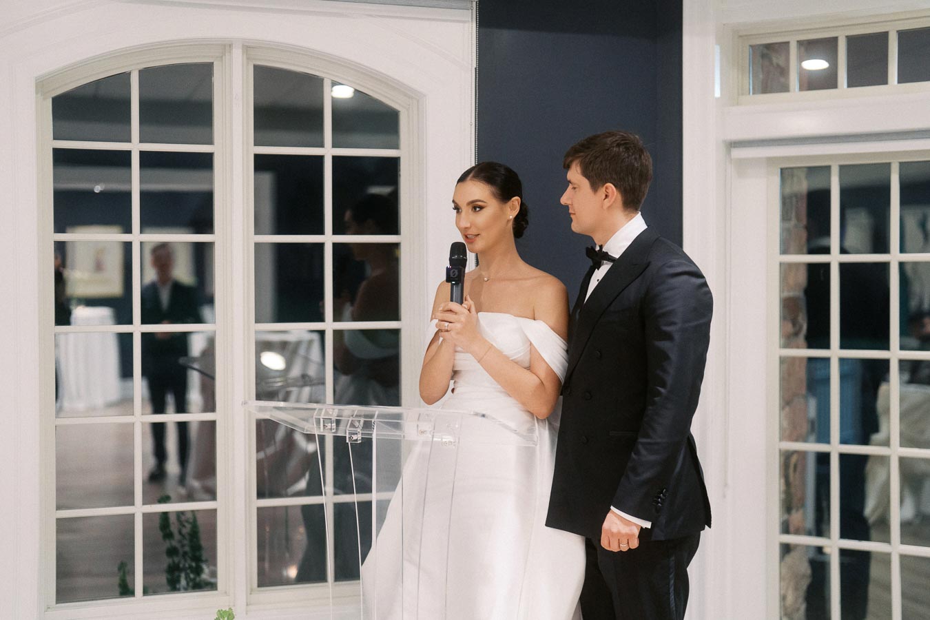 Bride and groom in elegant attire giving a speech at their wedding reception, standing at a transparent podium in front of large windows.