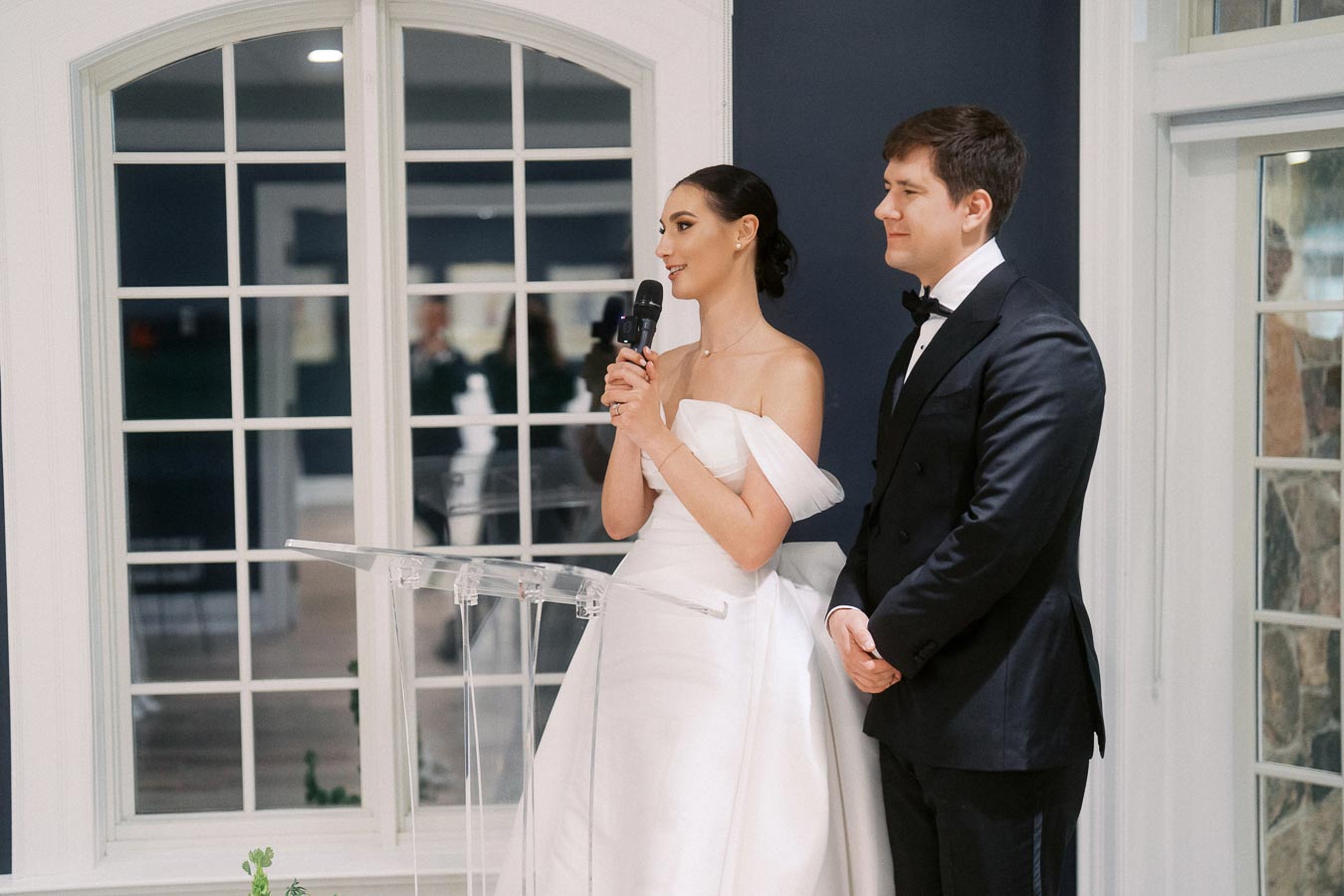 A bride and groom in formal attire stand together at a podium, with the bride holding a microphone, in a well-lit room with large windows.