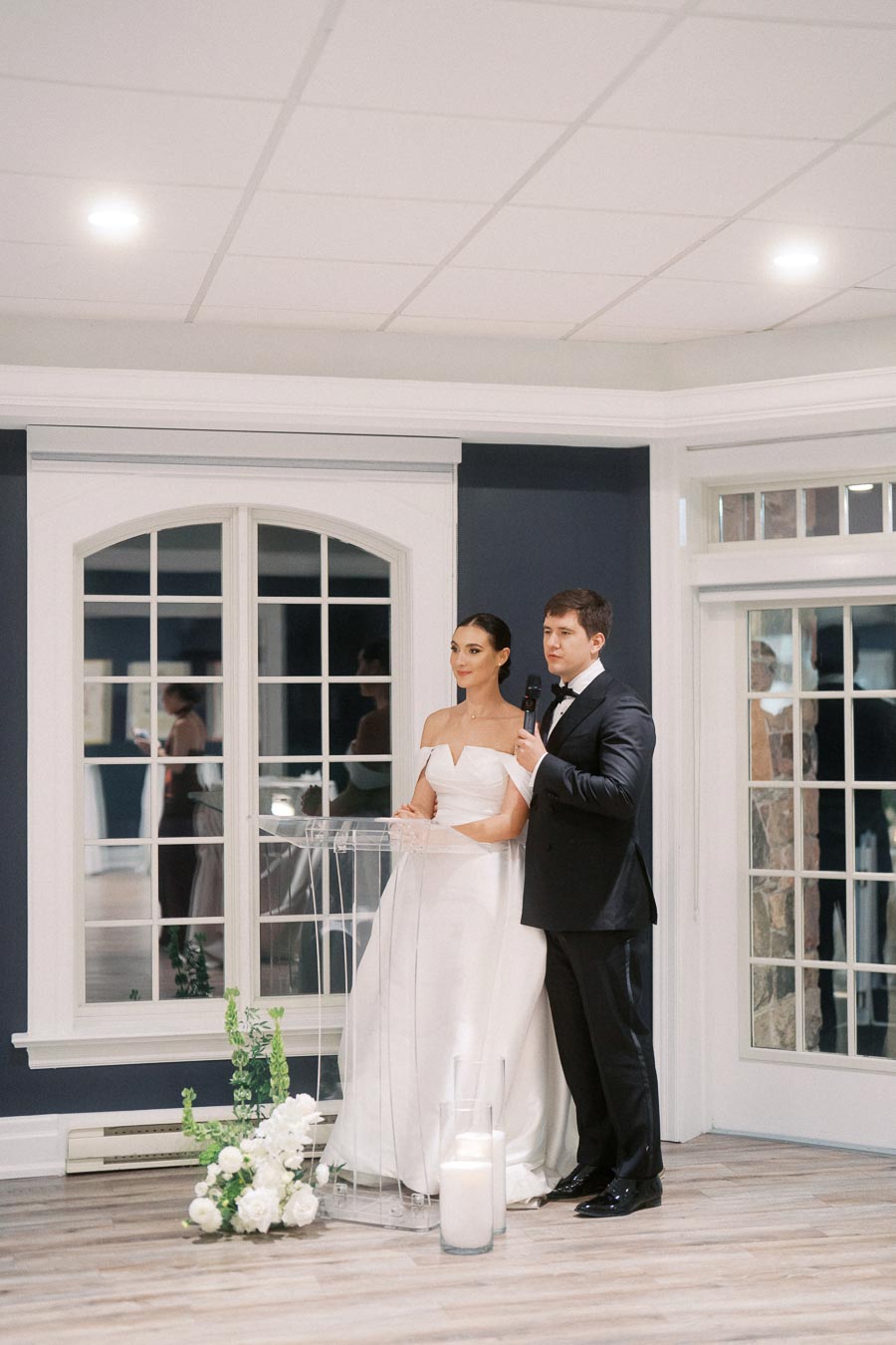A bride and groom give a speech indoors at their wedding reception. The bride wears an elegant white dress and the groom a classic black tuxedo. They stand beside a clear podium with floral decorations, framed by large windows and soft lighting.