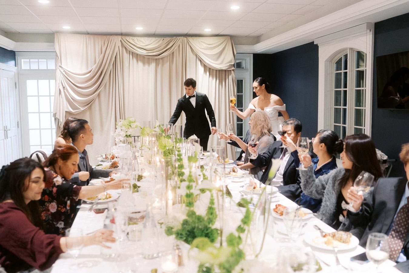 Elegant wedding dinner reception with guests toasting at a beautifully decorated long table adorned with floral arrangements and candlelight in a sophisticated dining room.