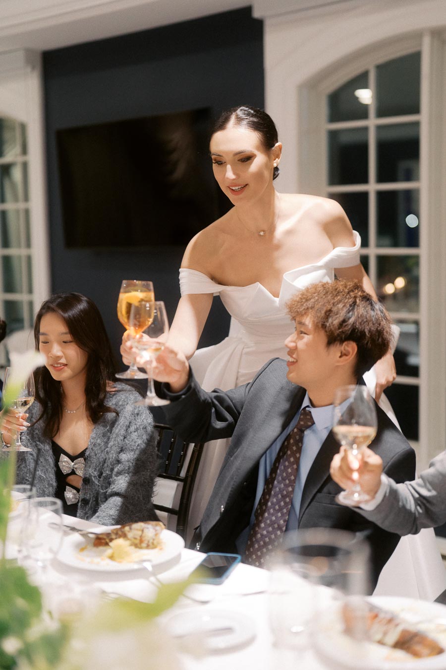 Elegant dinner party scene with guests raising glasses for a toast, featuring a woman in a white dress engaging with seated attendees in formal attire, set in a stylish and warmly lit dining room.